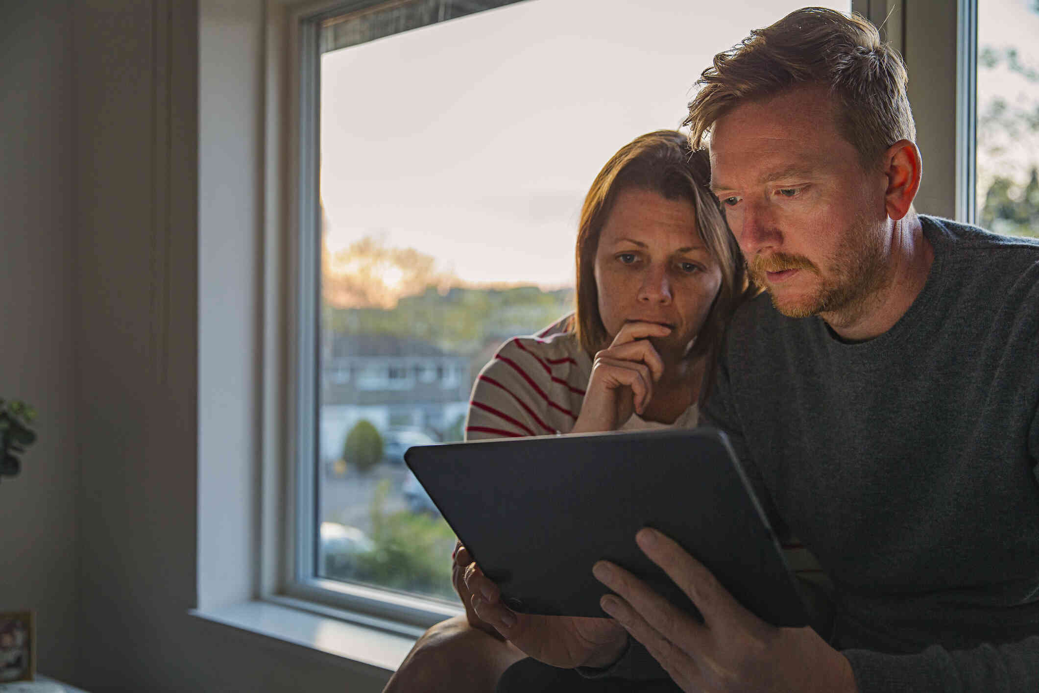 A woman with a worried expression covers her mouth with her hand as she sits by a window next to a man who is holding and looking down at a tablet.